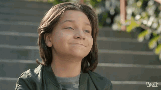 a young boy with long hair is smiling with the word brat behind him