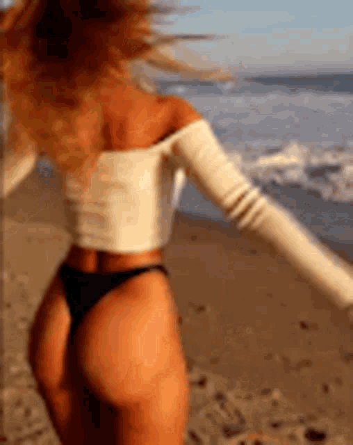a woman in a white top and black bikini is standing on a beach with her arms outstretched .