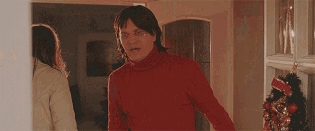 a man in a red turtleneck sweater stands in front of a christmas tree