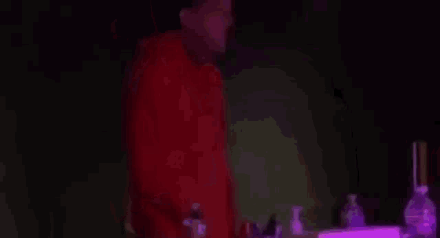 a man in a red shirt is standing in front of a table with bottles of alcohol .