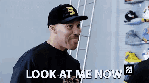 a man wearing a hat with the letter b on it is standing in front of a wall of shoes and says look at me now .