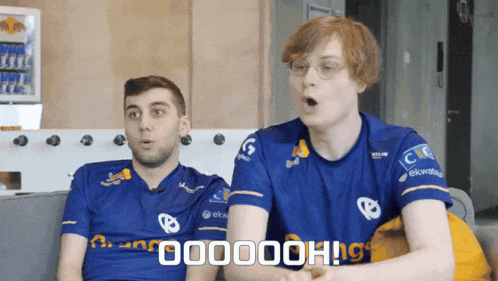 two men are sitting on a couch and one of them is wearing a blue shirt that says 000000h