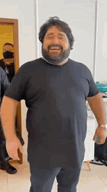 a man with a beard is wearing a black t-shirt and smiling while standing in a room .