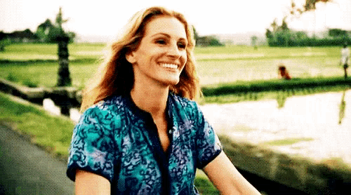 a woman in a blue shirt is smiling while riding a bike in a park .