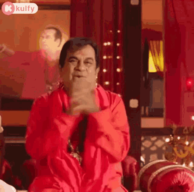 a man in a red shirt is sitting on a couch with his hands folded and making a funny face .
