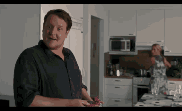 a man in a black shirt is standing in a kitchen with a woman behind him