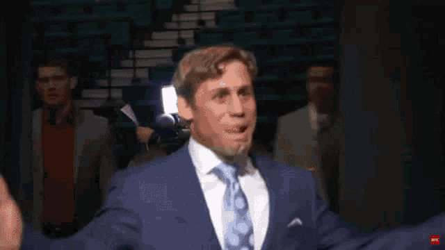a man in a blue suit and tie stands in a dark room with his arms outstretched