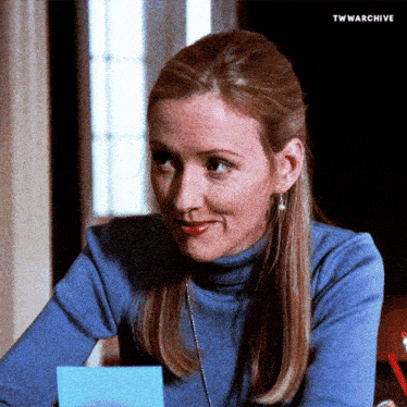 a woman in a blue turtleneck sits at a desk with a blue sticky note in her hand