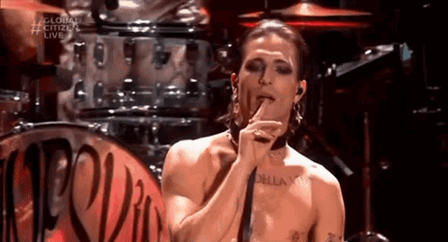 a shirtless man is singing into a microphone on a stage with a drum set in the background .