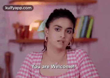 a woman in a pink checkered shirt is standing in front of a bookshelf and says `` you are welcome '' .