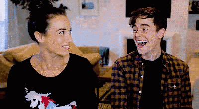 a man and a woman are laughing together in a living room . the man is wearing a unicorn sweater .