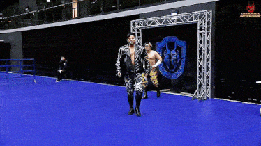 a man in a leather jacket walks on a blue mat in front of a dragon gate network sign