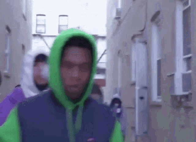 a man in a green hoodie is walking down a narrow street .