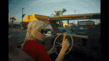 a woman wearing sunglasses is driving a car with a yellow building in the background