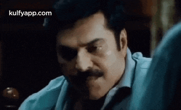 a man with a mustache is sitting at a table in a dark room and making a funny face .