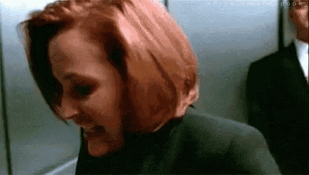 a woman with red hair is smiling in an elevator while a man in a suit stands behind her .