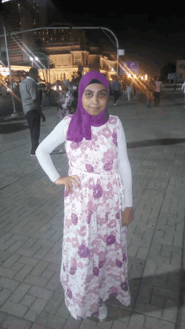 a woman wearing a purple hijab and a floral dress stands on a brick sidewalk