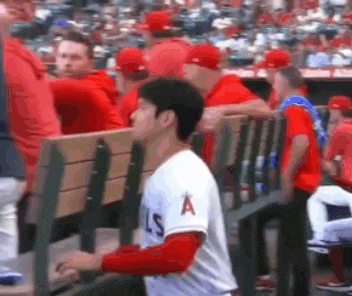 a baseball player is sitting in the dugout at a baseball game while a crowd watches .