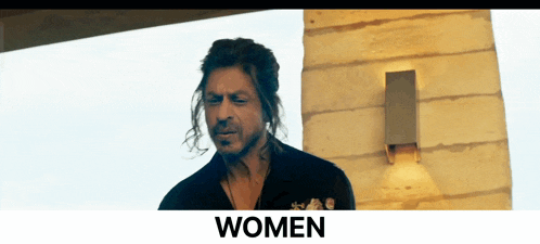 a man with long hair is standing in front of a wall with the word women written below him