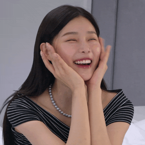 a woman wearing a pearl necklace and a striped shirt is smiling with her hands on her face