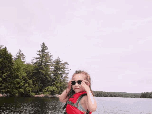 a little girl wearing sunglasses and a red life jacket stands on a lake