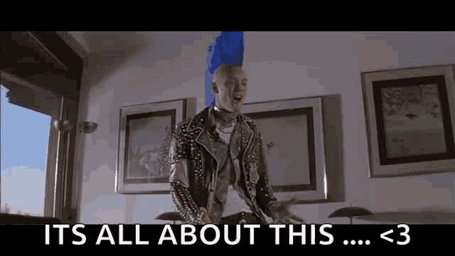a man with a mohawk is sitting on a couch and says it 's all about this .