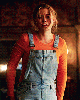 a woman wearing overalls and an orange shirt looks surprised