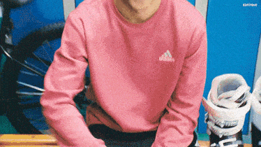 a man wearing a pink adidas sweatshirt is sitting on a bench next to a pair of ski boots .