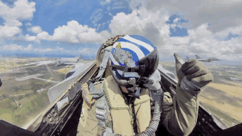 a pilot wearing a blue and white helmet with a greek flag on it gives a thumbs up