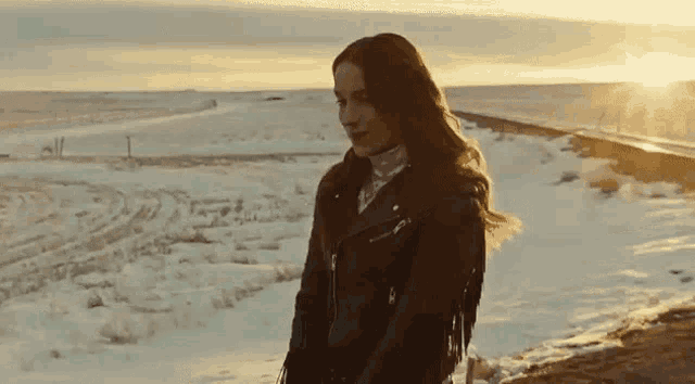 a woman in a leather jacket is standing in the snow near a body of water at sunset .