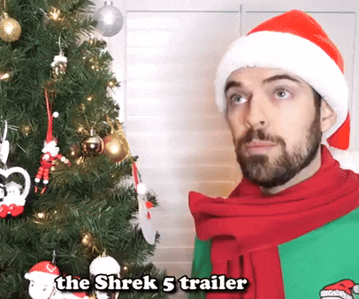 a man wearing a santa hat and scarf stands in front of a christmas tree with the words the shrek 5 trailer above him