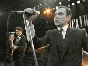 a man in a suit and tie is singing into a microphone on a stage while another man plays a guitar .