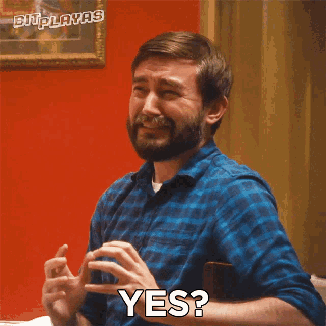 a man in a blue plaid shirt says yes in front of a red wall