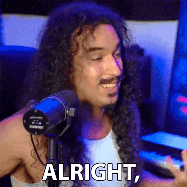 a man with long curly hair is standing in front of a microphone and says alright