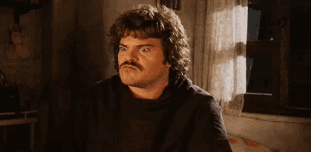 a man with a mustache is making a funny face while sitting in front of a window in a room .