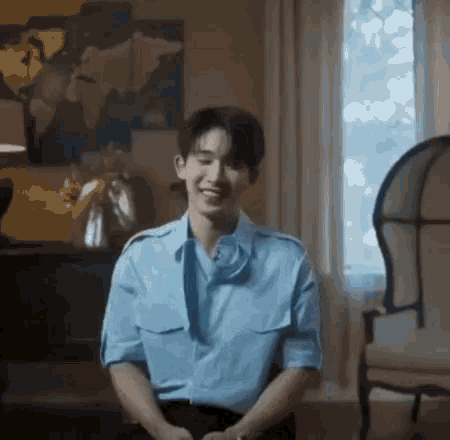 a young man in a blue shirt and tie is sitting in a chair in a living room and smiling .