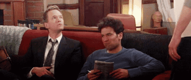 a man in a suit and tie is sitting on a couch next to another man .