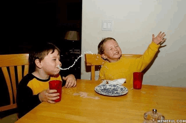 two young boys are sitting at a table drinking milk from red glasses while a plate of food is on the table