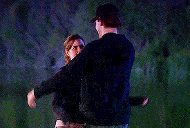 a man and a woman are hugging and looking at each other . the man is wearing a baseball cap .