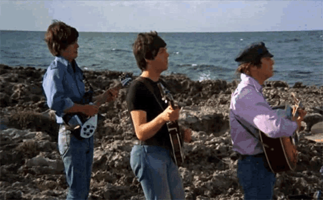 three men are playing guitars on a rocky beach