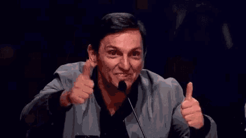 a man is giving two thumbs up in front of a microphone while sitting in front of a crowd .