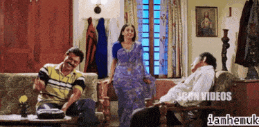 a woman in a purple sari is standing next to two men in a living room with a sign that says icon videos