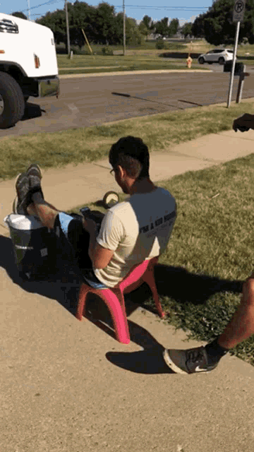 a man wearing a shirt that says " i 'm a kid again " sits on a pink chair on the sidewalk