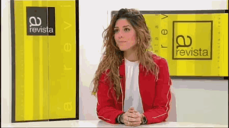 a woman in a red jacket sits at a desk in front of a revista sign