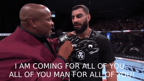 a man in a red suit is talking to a man in a black ufc shirt