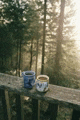 two cups of coffee are sitting on a wooden railing overlooking a forest .