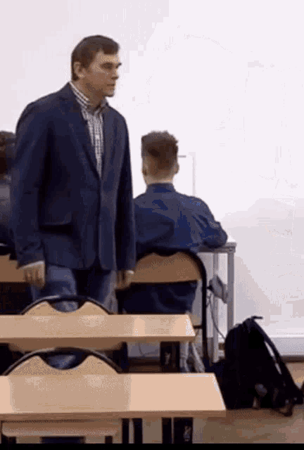 a man in a suit stands in a classroom talking to a student