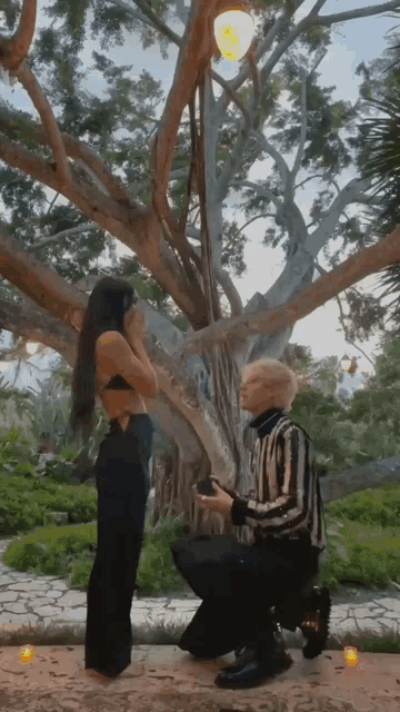 a man is kneeling down to propose to a woman in front of a tree