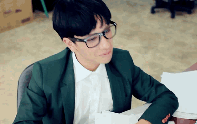 a man wearing glasses and a green jacket sits at a desk with papers