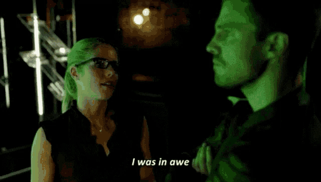 a man and a woman are standing next to each other in a dark room . the woman is wearing glasses .